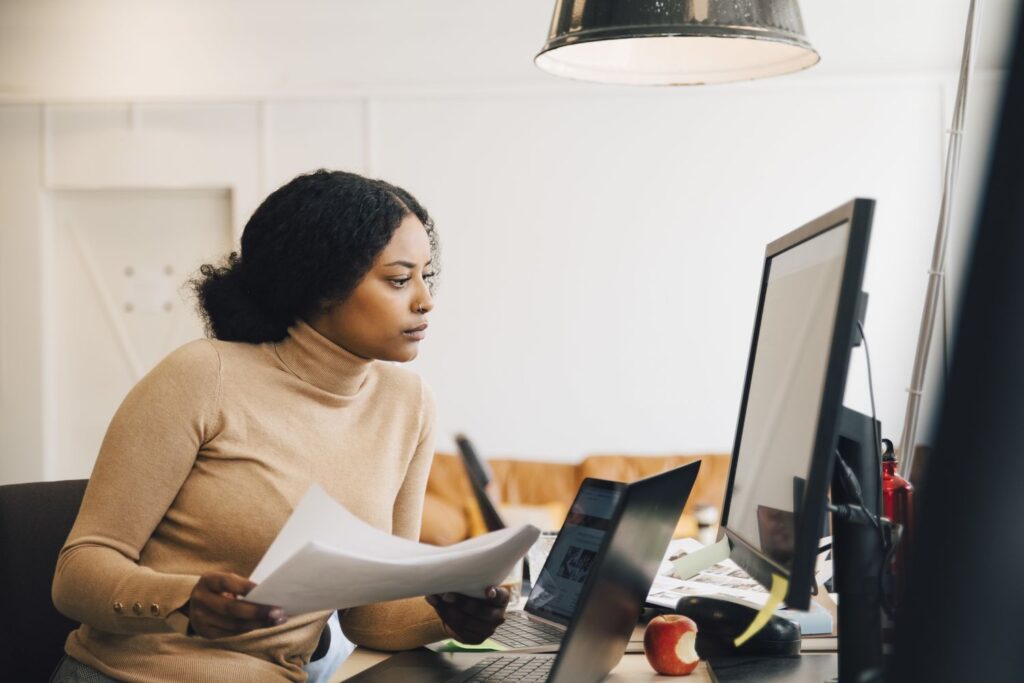 focused female it professional holding document while looking at computer in creative office 1171809